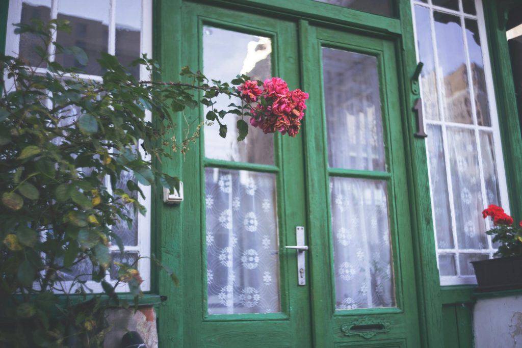 Close-up of a vintage green door adorned with vibrant pink flowers and lace curtains.