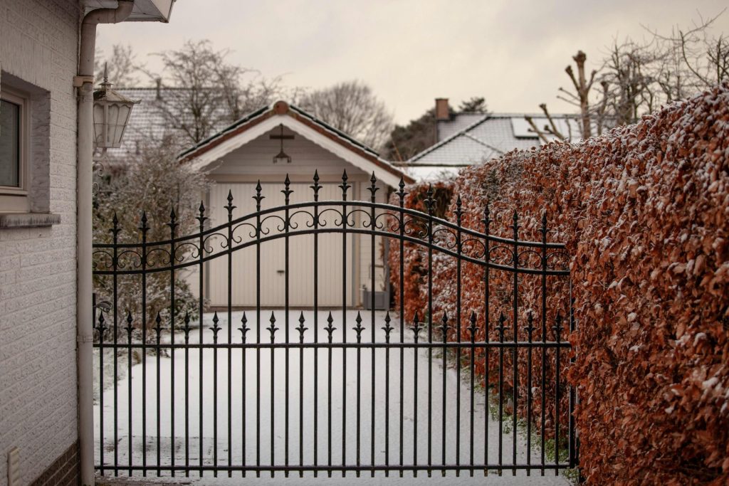 Une cour enneigée et sereine, ornée d'un portail en fer forgé et entourée de haies en hiver.