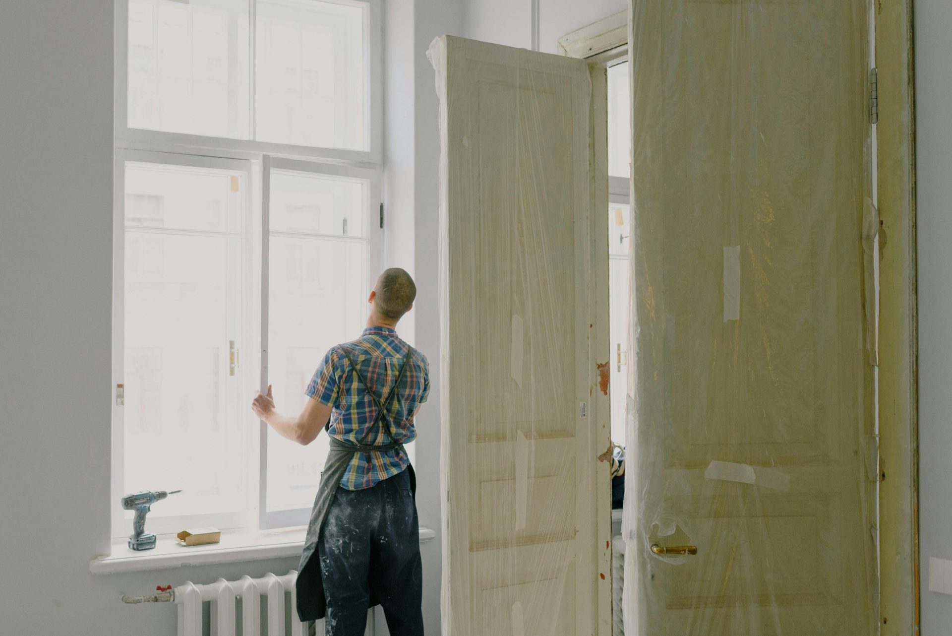 Vue de dos d'un ouvrier non identifiable en tablier, installant une fenêtre près d'une perceuse et de portes dans un appartement
